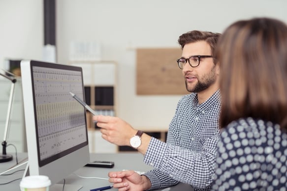 Proficient young male employee with eyeglasses and checkered shirt, explaining a business analysis displayed on the monitor of a desktop PC to his fem